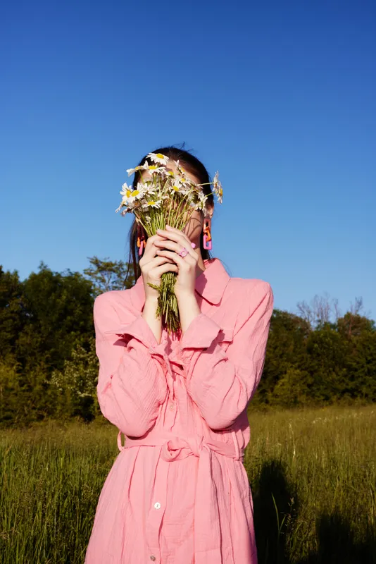 Young woman in a pink dress holding daisies in front of her face, standing in a sunny field, symbolizing joy and connection with nature