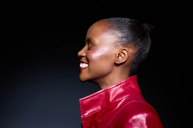 Profile of a smiling African woman with dark skin wearing a red leather jacket against a black background. Concept of joy, fashion, confidence, and beauty in studio lighting.