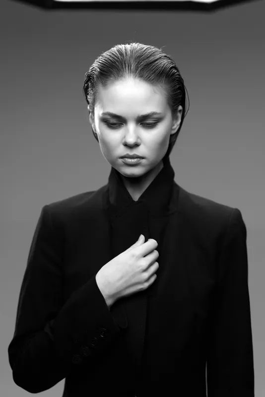 Black and white portrait of a serious woman in dark suit with contemplative expression and sleek hairstyle in studio lighting.