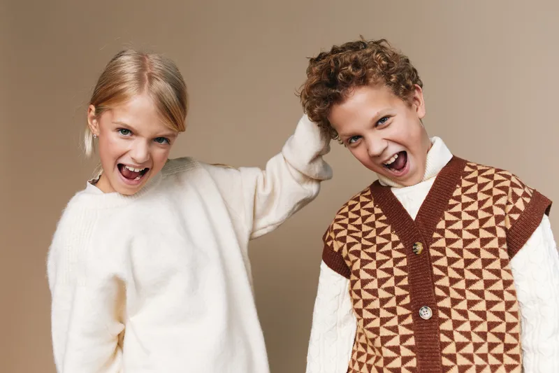 Two playful young models pose in a bright studio, laughing with energy as they touch hands, conveying friendship, fun, and spontaneous joy against a neutral backdrop.