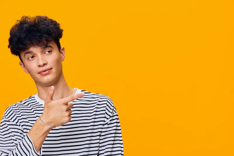 young man pointing finger with thoughtful expression wearing striped shirt against bright orange background, casual pose and modern hairstyle