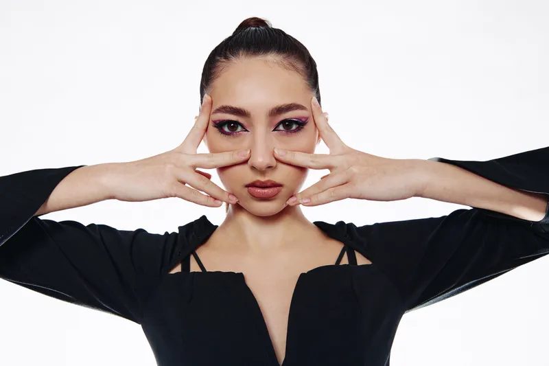 Young woman with artistic eye makeup poses with hands framing her face in expressive pose. Stylish black outfit creates bold fashion statement in creative beauty portrait.