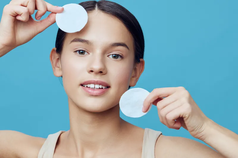 Young woman demonstrating skincare routine with cotton pads on a blue background, showcasing healthy skin and beauty concepts
