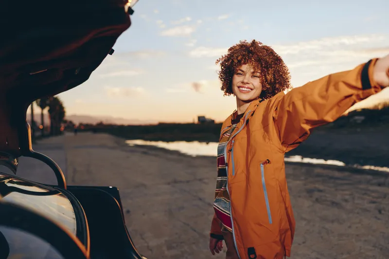 Smiling woman in an orange jacket meets someone at a car on a riverside walkway at sunset, casual lifestyle outdoors, travel mood, warm evening light, candid portrait