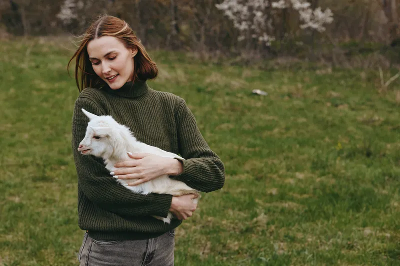 Young woman in a green sweater gently holds a white baby goat outdoors on a grassy field during a calm spring day.