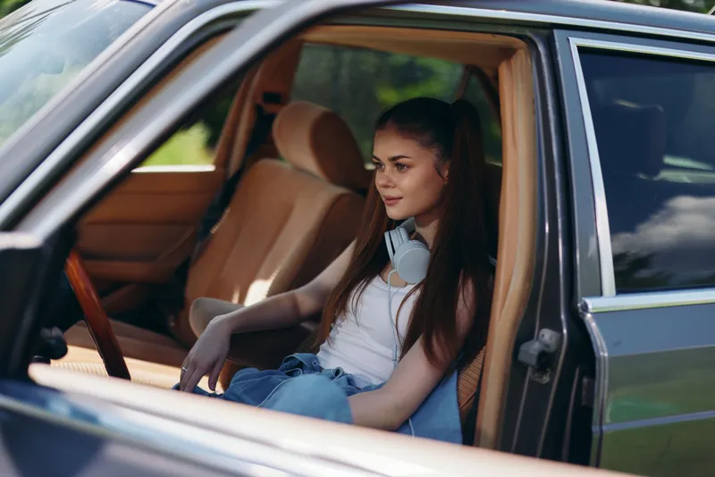 Young woman relaxing in a car, wearing casual clothing and headphones, reflecting feelings of peace and enjoyment during a sunny day.