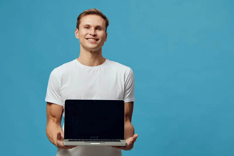 Smiling man holding open laptop computer with blank screen, casual white t-shirt, against plain blue background, studio shot, lifestyle concept, modern technology, young adult male, happy expression, 