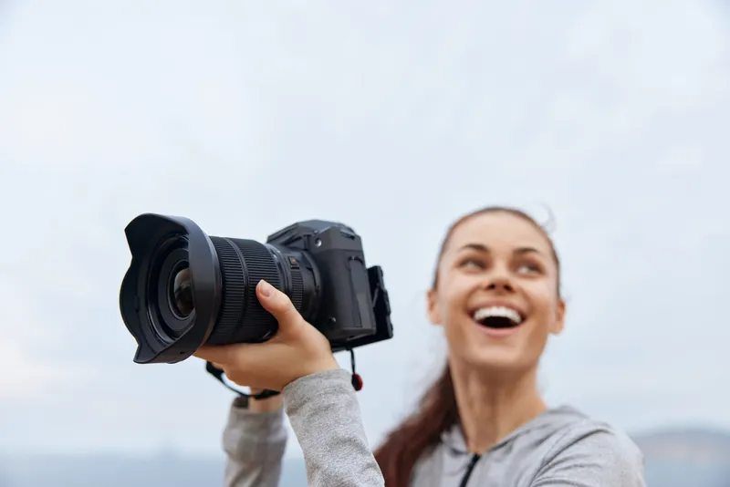 Young woman with camera smiling outdoors, showcasing passion for photography, fashion in activewear, capturing moments against a scenic background