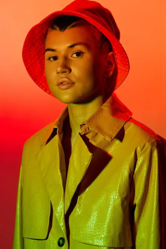 young woman wearing a red bucket hat and light yellow leather jacket with confident expression against vibrant orange and red gradient background, modern portrait style