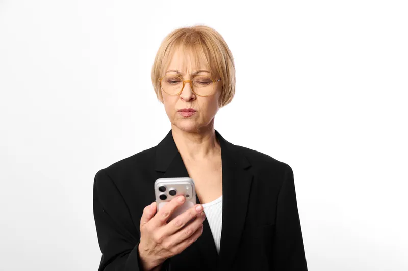 Woman senior holding smartphone and glasses indoors, technology communication, looking down for texting, reading on screen, expression, white background