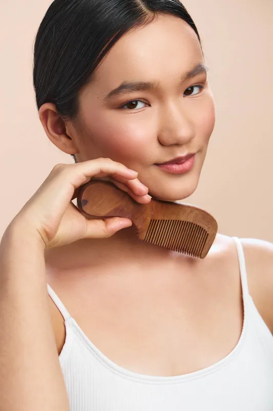 Asian woman holds a wooden comb near her face in a studio portrait. She gazes softly at the camera with a calm smile, conveying natural beauty and mindful grooming.