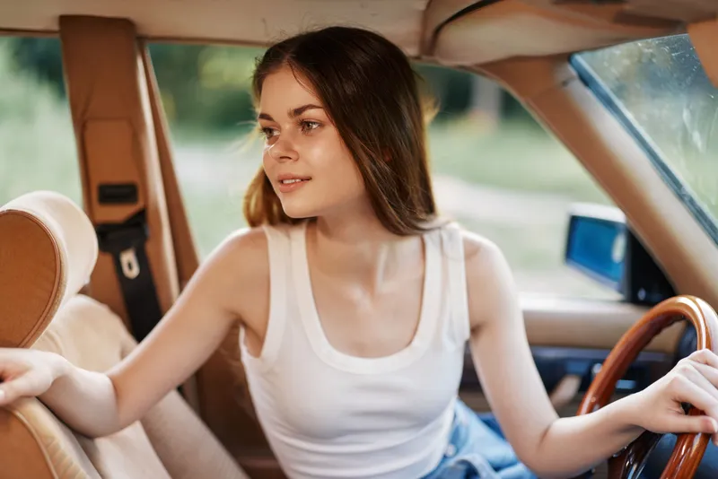 Young woman in a sleeveless top enjoying a drive in a classic car, exuding a relaxed and cheerful vibe amidst a natural green background.