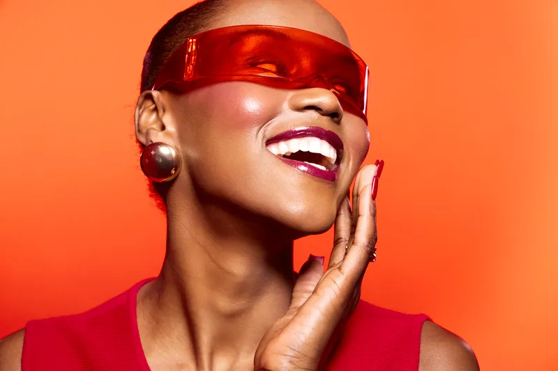 Smiling black woman with dark skin wearing futuristic red visor and bold earrings poses against orange background. Concept of fashion, beauty, confidence, and modern style.