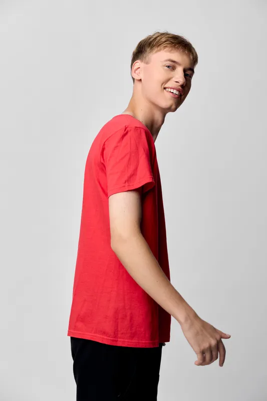 Youthful man wearing a red shirt in a casual pose, smiling at the camera in a clean studio setting, lifestyle fashion vibe, relaxed mood and approachable style.