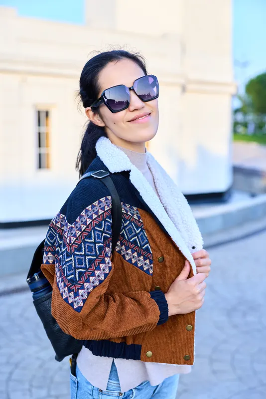 Woman wearing a patterned sleeve jacket, scarf and sunglasses posing outdoors with a backpack, showcasing casual street fashion, confident pose and sunny style.