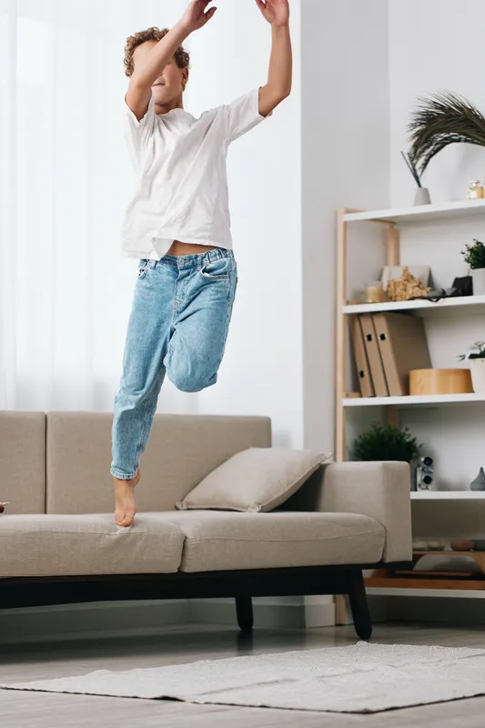 Energetic person mid jump on a cozy living room sofa, captured in bright natural light, expressing joy, spontaneity, and carefree movement.