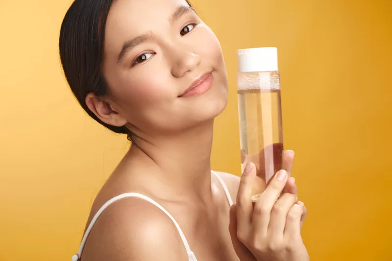 asian woman smiles softly while holding a clear bottle of liquid skincare product in a warm orange studio, conveying a fresh, confident mood and beauty routine.