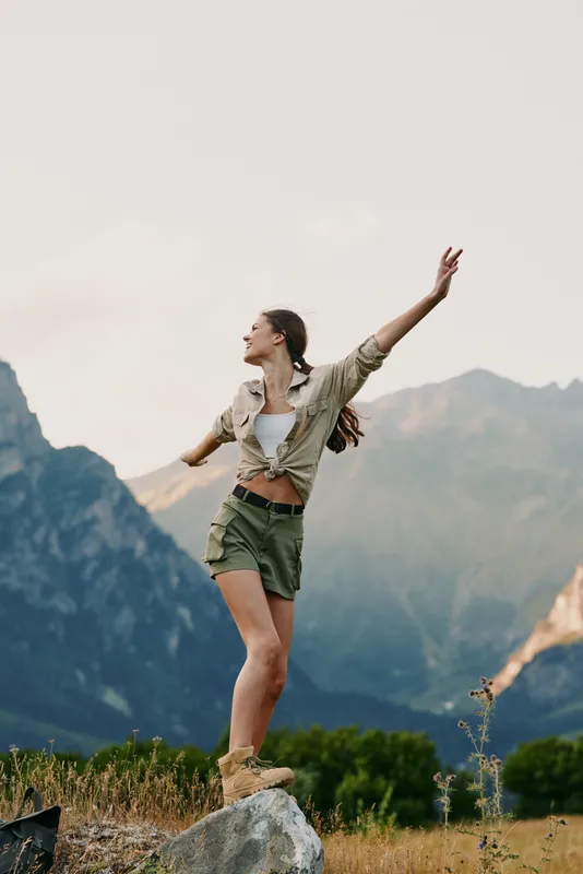 Young woman stands on a rocky outcrop in a mountain landscape with arms raised, enjoying the breeze and the sense of freedom