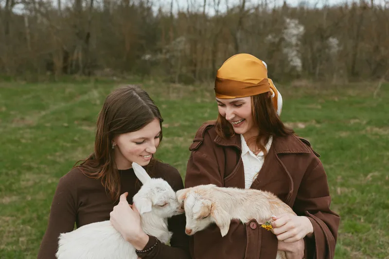 Two women share a warm moment with playful goats in a sunny meadow, showcasing casual countryside lifestyle and friendly animal interaction, joyful smiles as they cradle the kid goats