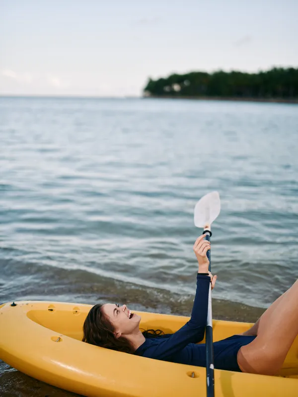 A person relaxes in a yellow kayak on calm coastal water, paddling with a raised paddle while distant shoreline and clear sky create a peaceful outdoor scene.