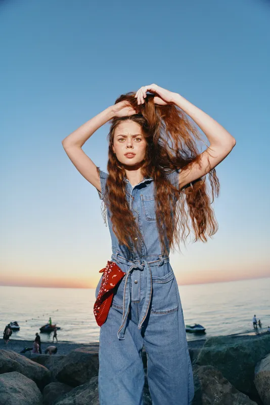 young woman in denim jumpsuit stands by the sea at sunset with long hair styled confidently and a red scarf belt accessory