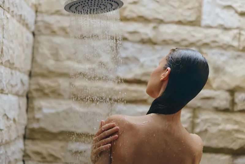 Woman taking shower under rainfall showerhead with stone wall background, wet hair, and relaxed posture in calm bathroom setting.