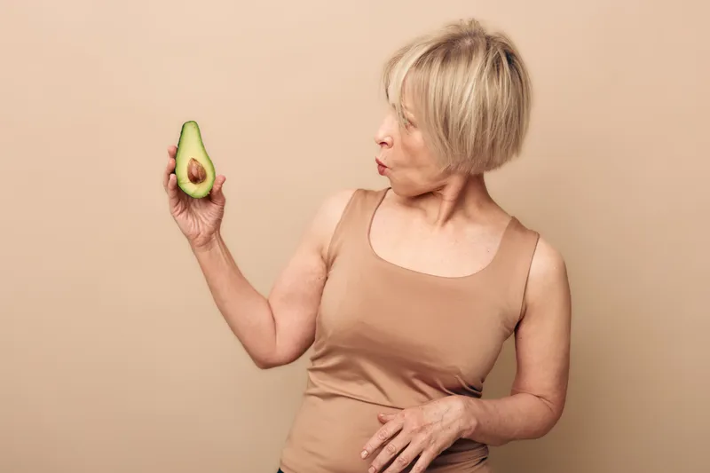Mature woman with short blonde hair holding half an avocado looks surprised. Beige background and casual sleeveless top create a natural, healthy lifestyle concept.