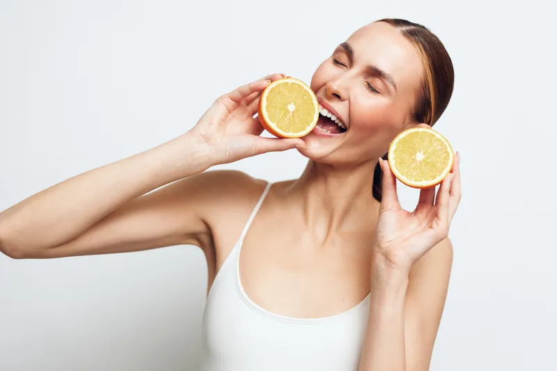 Young woman with closed eyes holding two halves of a fresh orange near her face, smiling joyfully, natural light, clean white background, healthy lifestyle concept.