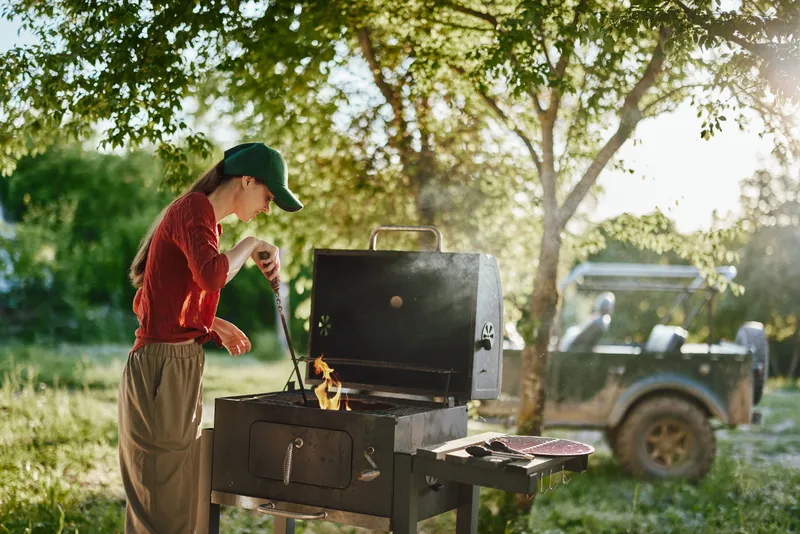 Young woman grilling outdoors in a sunny garden, wearing a red shirt and green cap, showcasing summer cooking lifestyle and joy of outdoor BBQ gatherings