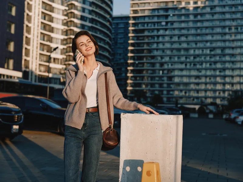 A cheerful woman speaks on a mobile phone on an urban street. She smiles warmly, standing near a concrete barrier with tall buildings glowing in the background.