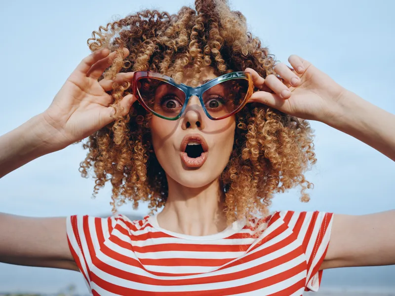 Vibrant portrait of a surprised woman with curly hair wearing colorful cat-eye sunglasses and a red white striped shirt, set against a clear blue sky.
