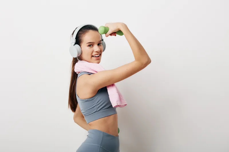Young athletic woman in gray sportswear with white headphones and pink towel, performing a bicep curl with a green dumbbell against a clean white studio background, smiling.