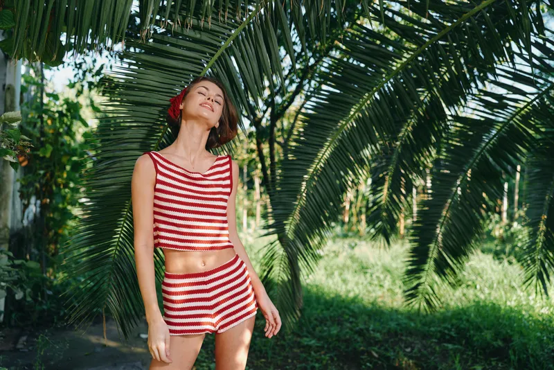 A cheerful young woman in a red and white striped bikini stands outdoors among lush palm trees, enjoying a sunny day and a relaxed, tropical vibe.
