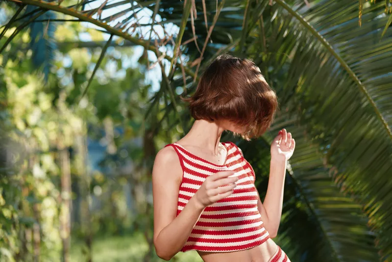 A cheerful young woman wearing a striped croptop enjoys sunshine outdoors. She smiles softly, moves gracefully, expressing carefree style and playful mood amid lush garden setting.