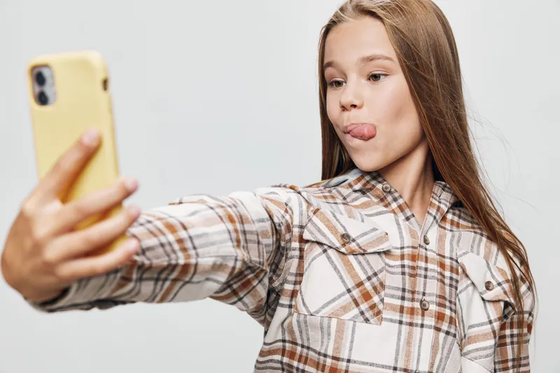 Teenage girl taking selfie with smartphone and making funny face sticking out tongue. Casual clothing, long hair, isolated on light background for social media content or communication.