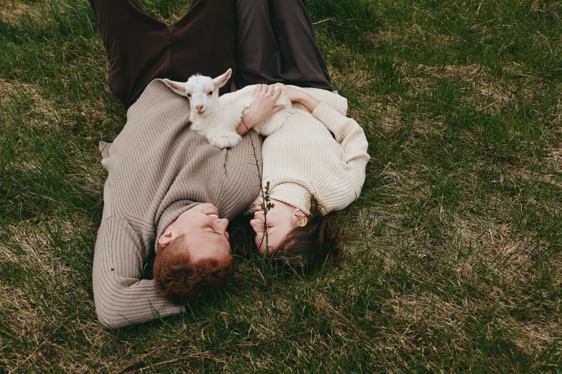 A young couple lies on the grass holding a small white baby goat while wearing cozy knitted sweaters in a natural outdoor setting, capturing a calm and intimate lifestyle moment.