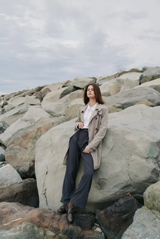 Young woman in casual outfit stands against large rocks outdoors on cloudy day. Fashion concept with natural background and relaxed confident pose by the seaside landscape.