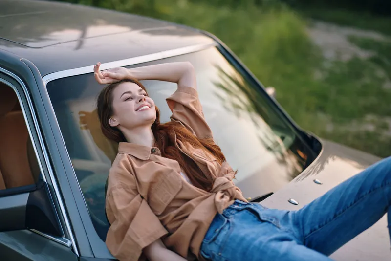 Young woman enjoying a sunny day, relaxing on a vintage car with a carefree smile, dressed in casual beige outfit and denim jeans, encapsulating summer vibes.