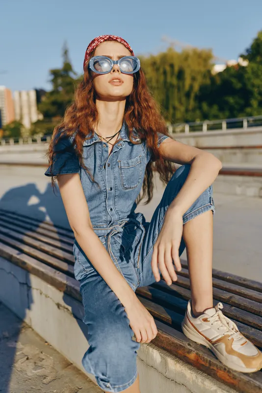 Lifestyle woman wearing modern boho-western denim street style outfit with sunglasses sitting outdoors in warm natural light, film color effect enhancing relaxed vibe.