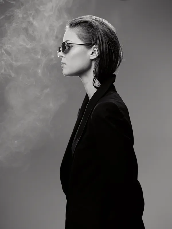 Black and white portrait of a stylish woman with sunglasses and slicked-back hair exhaling smoke in a dark suit.