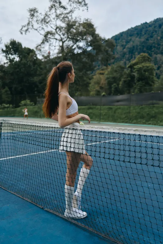 Young athletic woman in sporty attire, enjoying a moment on a tennis court surrounded by nature, showcasing style and confidence during afternoon practice