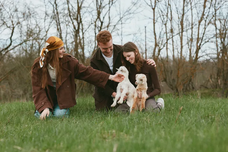 Happy family outdoors with a playful goat kid on a grassy field, children and adults smile while interacting with the animals, enjoying a wholesome countryside moment in daylight.
