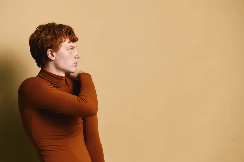 Portrait of young man with red hair wearing brown turtleneck standing against beige wall looking sideways with serious expression in studio lighting. Fashion and casual style concept.