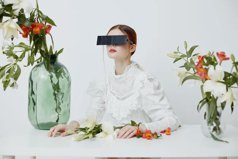 A fashion model with a blindfold styled in a white dress, surrounded by vibrant flowers, conveying a sense of mystery and beauty in a minimalistic setting