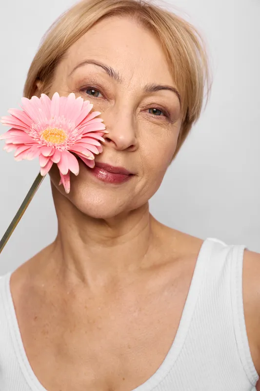 Woman portrait with gerbera flower in hand, mature face in soft light, pink petals by lips, calm smile, skincare concept, white background, elegance, femininity, relaxation