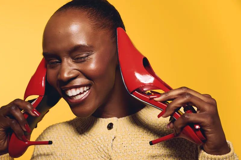 Happy African woman with dark skin holds red high heels near her ears, smiling and winking. Yellow background and warm lighting create joyful and playful mood for fashion concept.