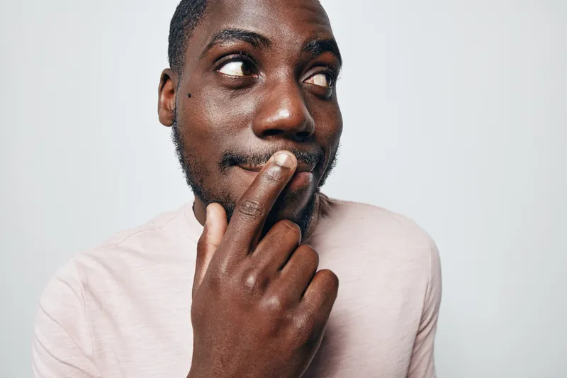 African American man with dark skin wearing light pink shirt looking sideways with thoughtful expression and finger on lips isolated on white background.