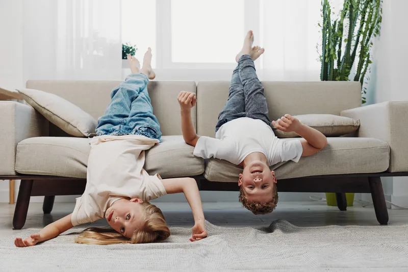 Two happy children lying upside down on sofa in bright living room, playful mood and fun family moment indoors at home.