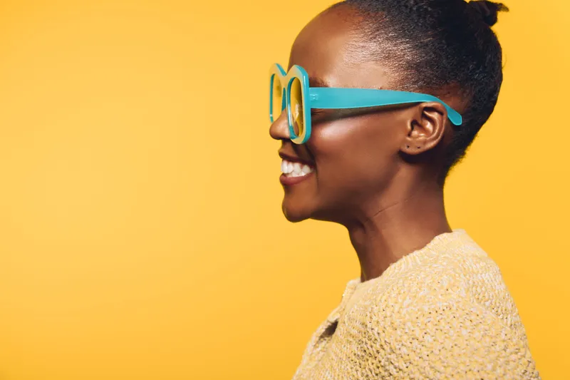 Profile of smiling dark skinned African woman wearing blue oversized glasses and yellow knitted sweater on bright yellow background. Concept of happiness, fashion, and positive mood.