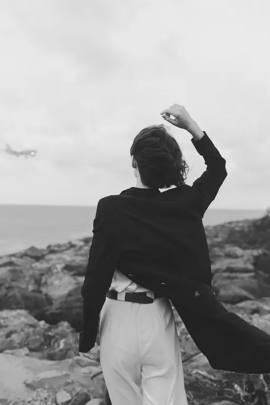 Coast figure, back view, wind in the coat, rocky shoreline and horizon capture freedom and exploration in a dramatic monochrome outdoor landscape with misty air.